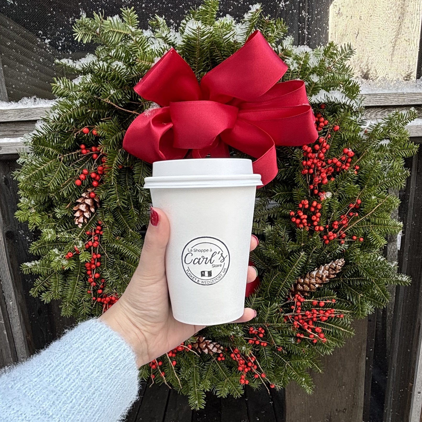 A hand holds a disposable coffee cup with La Shoppe à Carl / Carl's Store branding in front of a holiday wreath while snow falls.